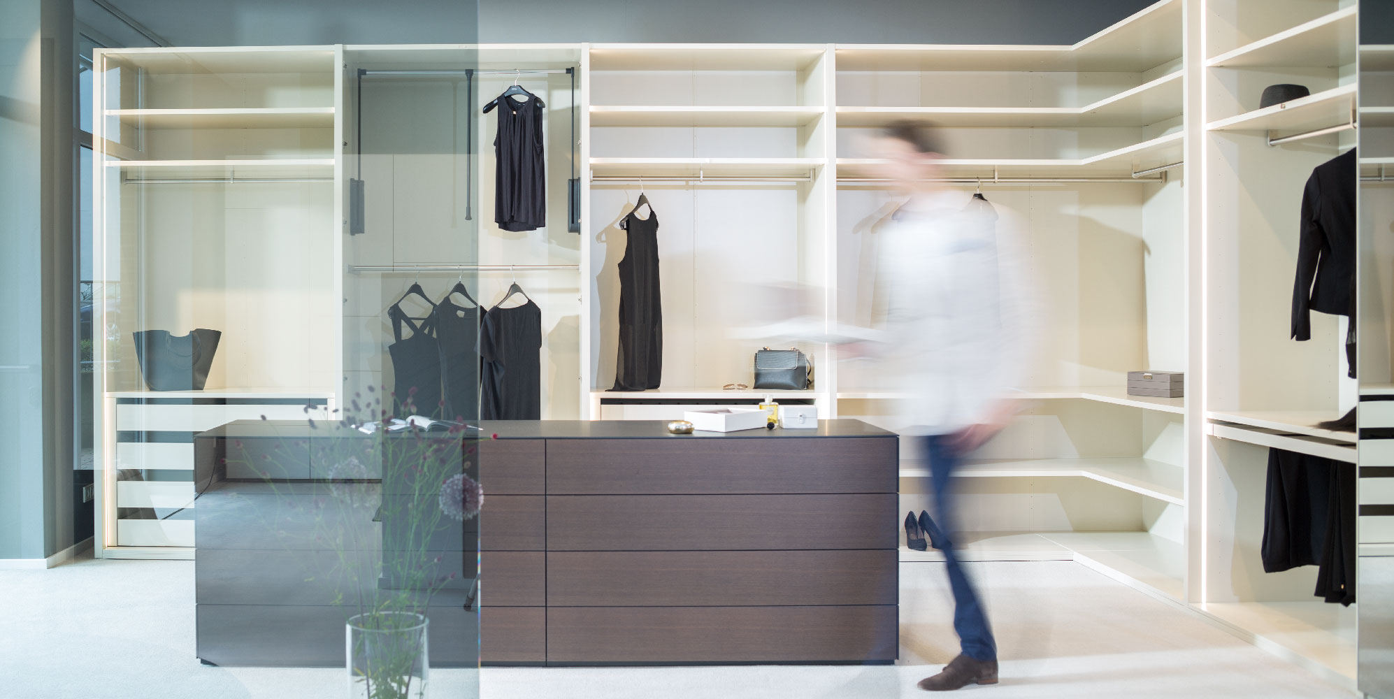 Begehbarer Kleiderschrank mit hellen Regalen und dunkler Mittelkommode bei Schründer Schlafräume.