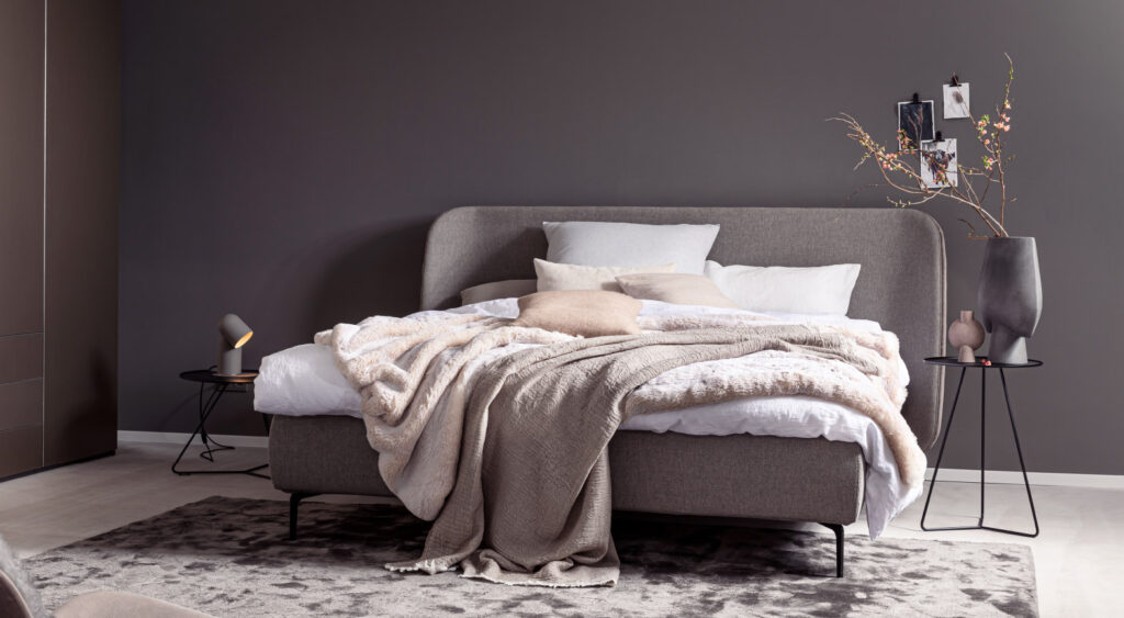 Ein stilvoll eingerichtetes Schlafzimmer mit Bett, Teppich und Beistelltischen in warmen Grau- und Pudertönen.