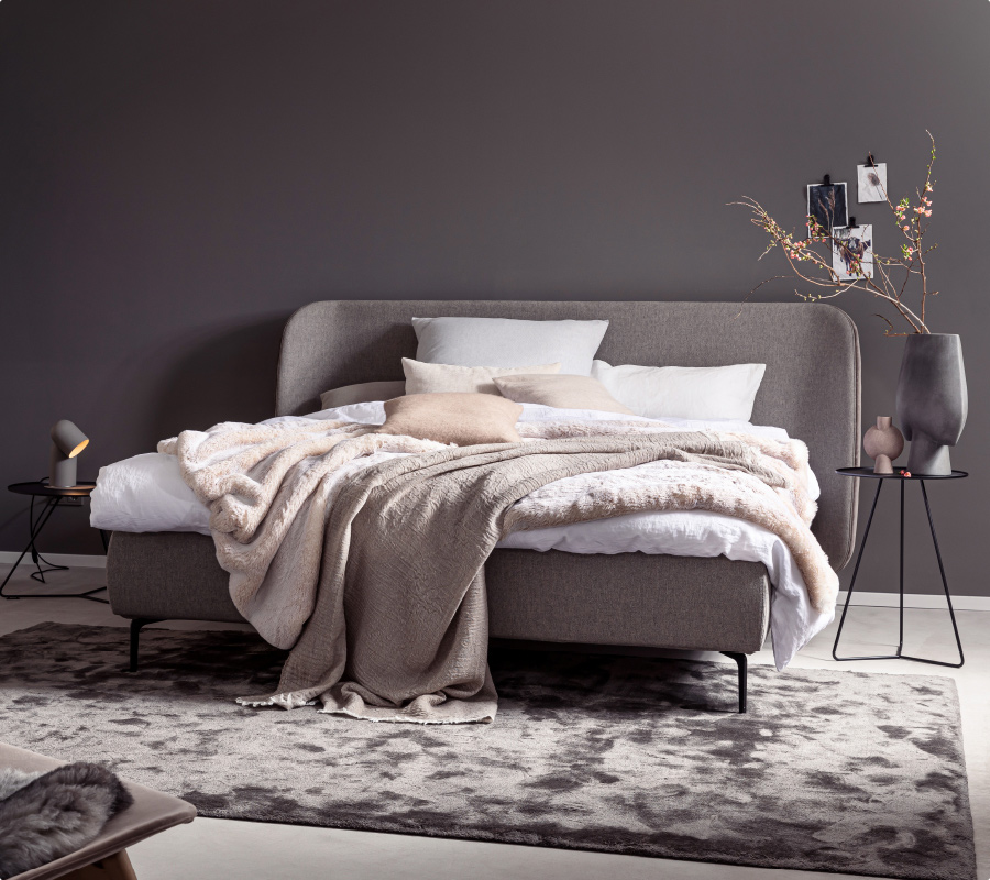 Ein stilvoll eingerichtetes Schlafzimmer mit Bett, Teppich und Beistelltischen in warmen Grau- und Pudertönen.