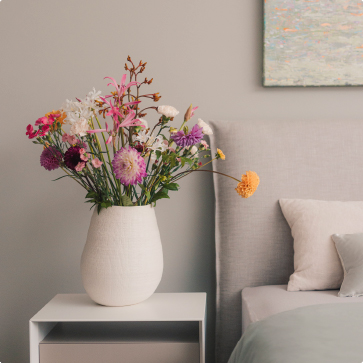Eine Vase mit einem bunten Blumenstrauß steht auf einem Designer-Nachtisch neben einem hochwertigen Bett.