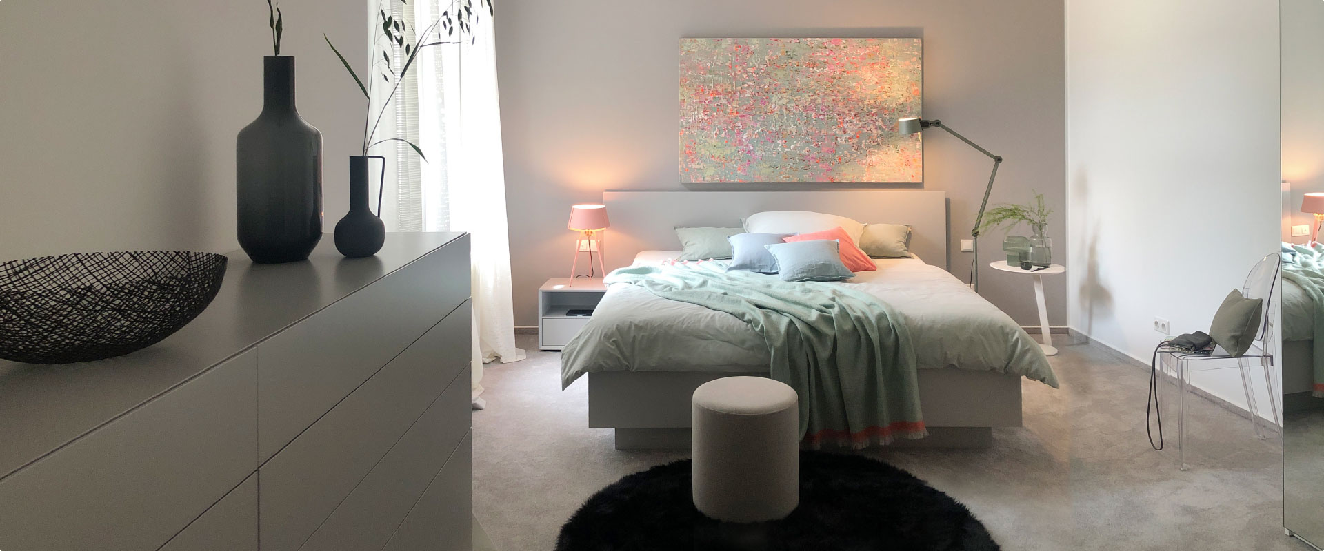Ein stilvoll eingerichteter Schlafraum mit Bett, Kleiderschrank und Beistelltisch in Grautönen und Farbakzenten in Koralle und Salbei.