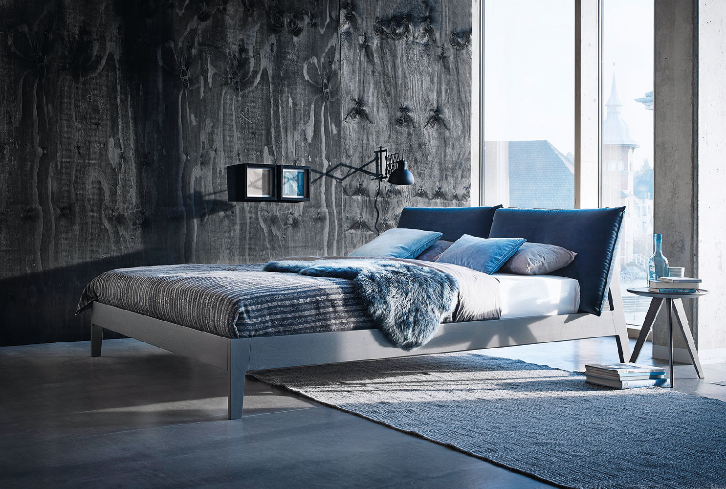 Modernes Designerbett vor dunkler Strukturwand mit großen Fenstern und minimalistischer Einrichtung