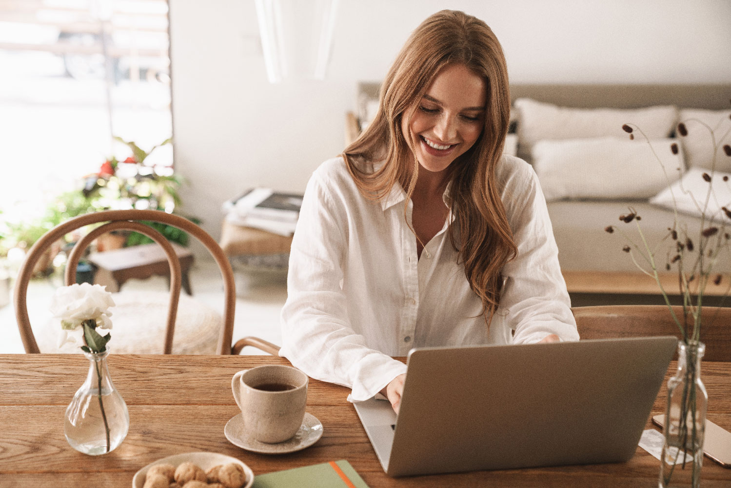 Junge Frau arbeitet lächelnd im Homeoffice am Laptop, sitzt an einem Holztisch mit einer Tasse Kaffee.