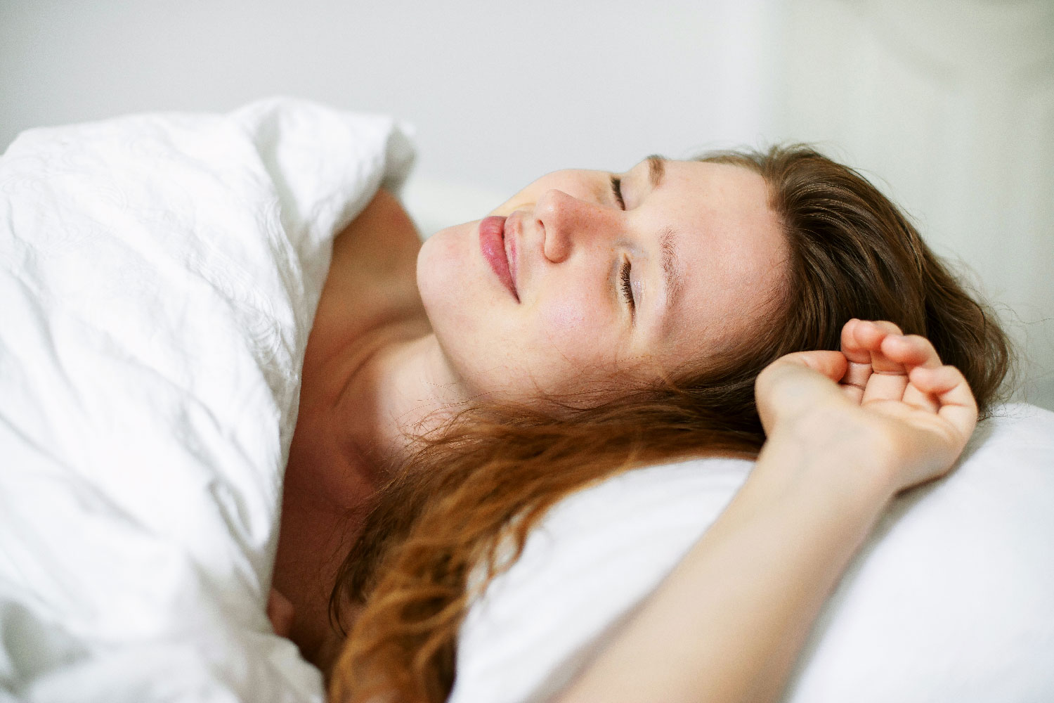Junge Frau liegt lächelnd und entspannt unter einer weißen Bettdecke mit geschlossenen Augen im Bett.