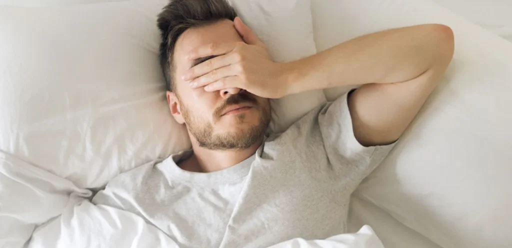 Mann im Tshirt liegt in einem weißen Bett auf dem Rücken und hält sich erschöpft die Hand über die Augen – Symbolbild für Schlafprobleme durch Schnarchen.